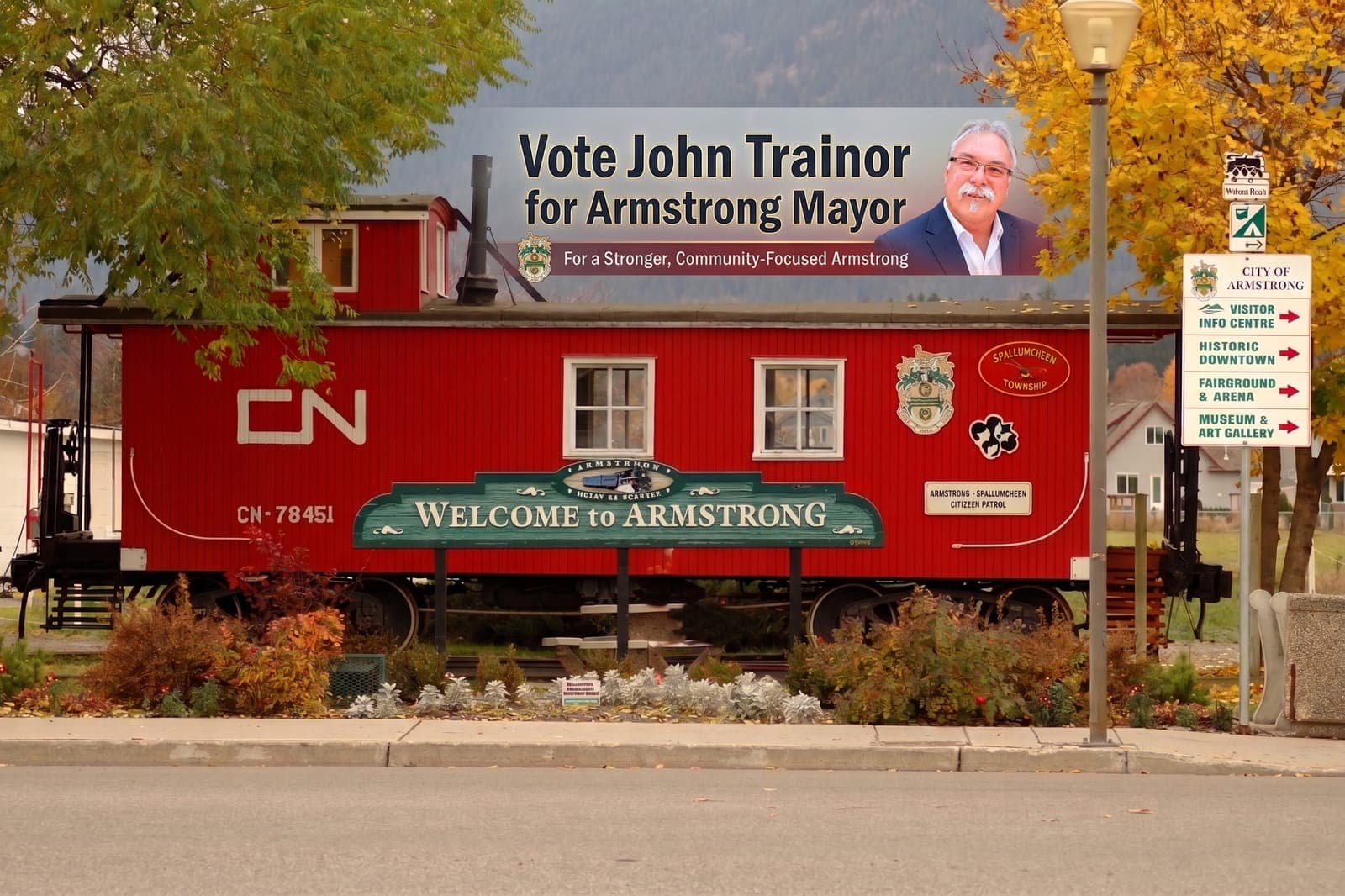 A "Vote John Trainor for Armstrong Mayor — For a Stronger, Community-Focused Armstrong" banner mounted atop a rail car next to the Welcome to Armstrong sign